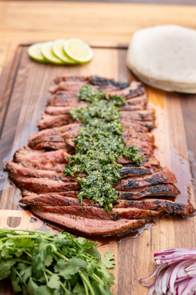Sliced smoked flank steak topped with cilantro chimichurri, served on a wooden board with tortillas, lime slices, fresh cilantro, and red onion—perfect for tacos.
