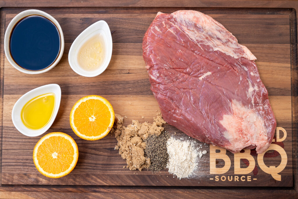 Raw flank steak on a wooden board surrounded by marinade ingredients: soy sauce, vinegar, olive oil, fresh orange halves, brown sugar, black pepper, and garlic powder—ready for smoking prep.