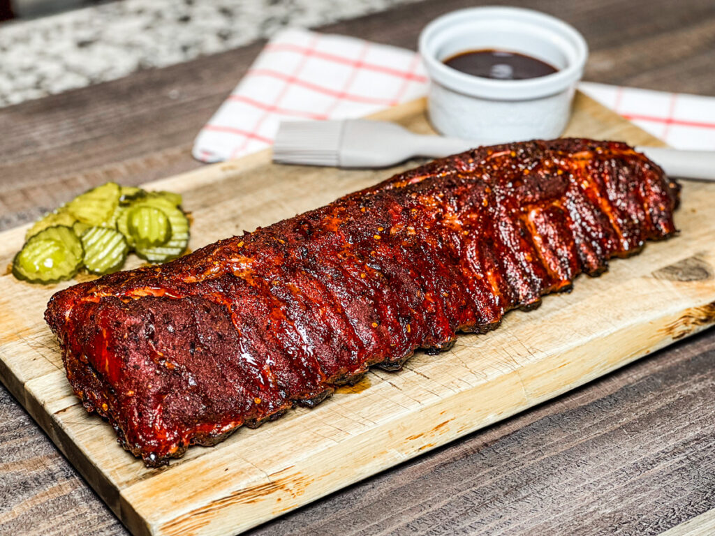 Rack of smoked baby back ribs with a rich mahogany bark, served on a wooden board with sliced pickles and a ramekin of barbecue sauce in the background.