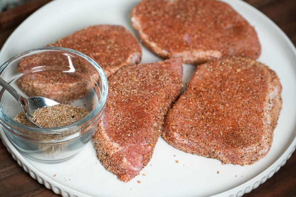 four raw seasoned pork chops on a white plate