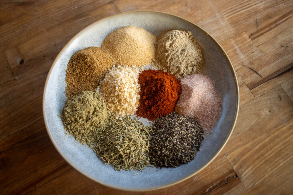A bowl filled with an assortment of vibrant spices and seasonings, including paprika, black pepper, garlic granules, rosemary, and pink salt, neatly arranged for a homemade smoked turkey rub on a wooden surface.