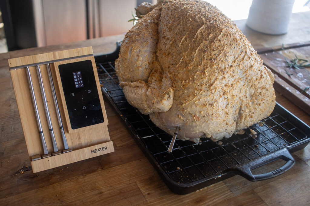 A seasoned turkey resting on a wire rack, prepped for cooking, with a MEATER thermometer set displayed nearby, showcasing its precision tools for monitoring the perfect cooking temperature.