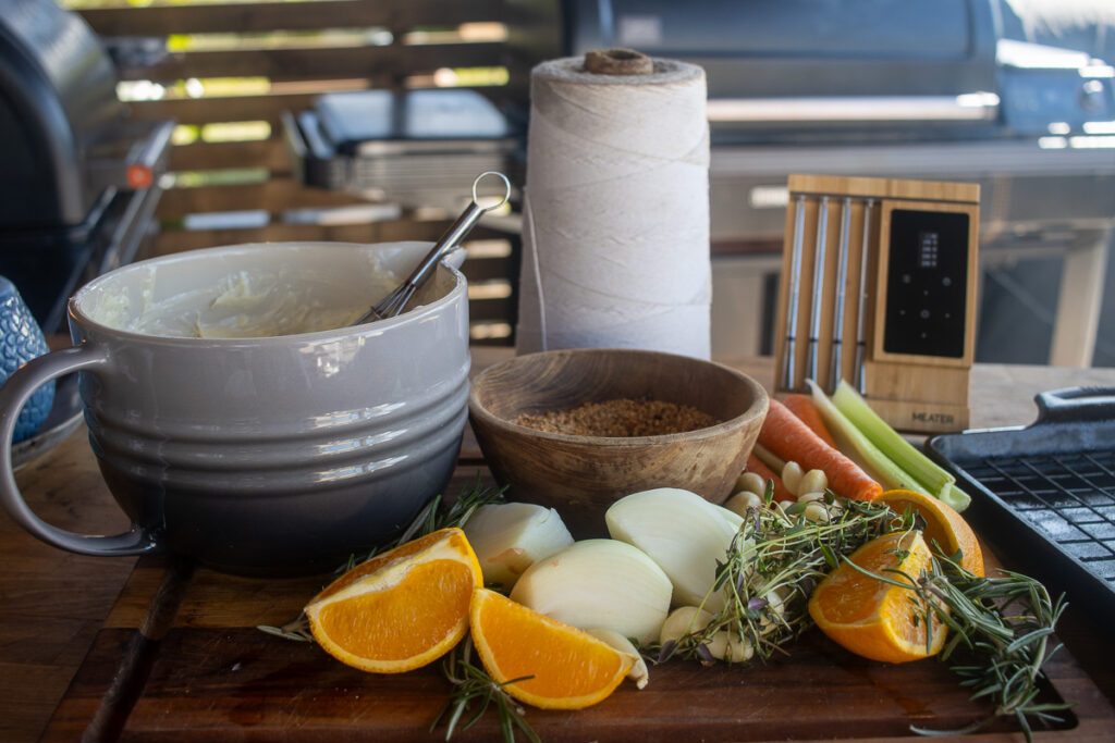 A rustic preparation scene featuring turkey ingredients: a bowl of seasoned compound butter, orange wedges, onions, garlic, carrots, celery, fresh rosemary, a bowl of spices, butcher's twine, and a MEATER thermometer, all set up for a pellet grill turkey recipe.