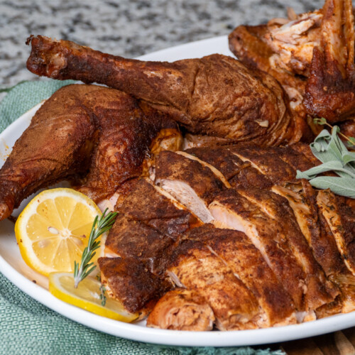 Smoked Cajun turkey carved and served on a white platter with lemon slices, fresh rosemary, and sage garnish, placed on a green napkin on a wooden surface.