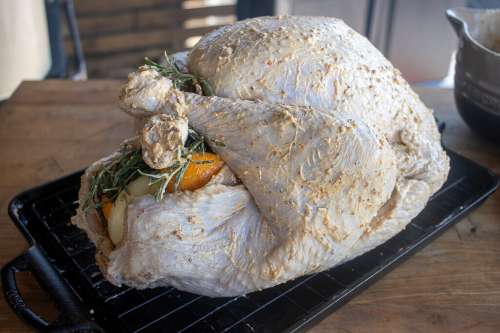 A whole turkey generously coated in a seasoned butter rub, stuffed with fresh orange slices, onions, and sprigs of rosemary, resting on a wire rack and ready for smoking.