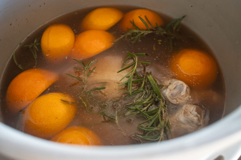 A turkey submerged in a flavorful brine inside a bucket, surrounded by halved oranges, fresh sprigs of rosemary, and rich, aromatic liquid, preparing for a perfectly seasoned dish.