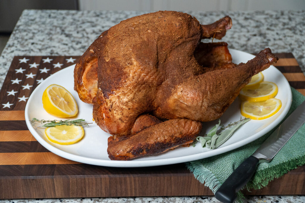 Whole smoked Cajun turkey served on a white platter with lemon slices, fresh thyme, and sage garnish, placed on a cutting board with an American flag design and a green napkin