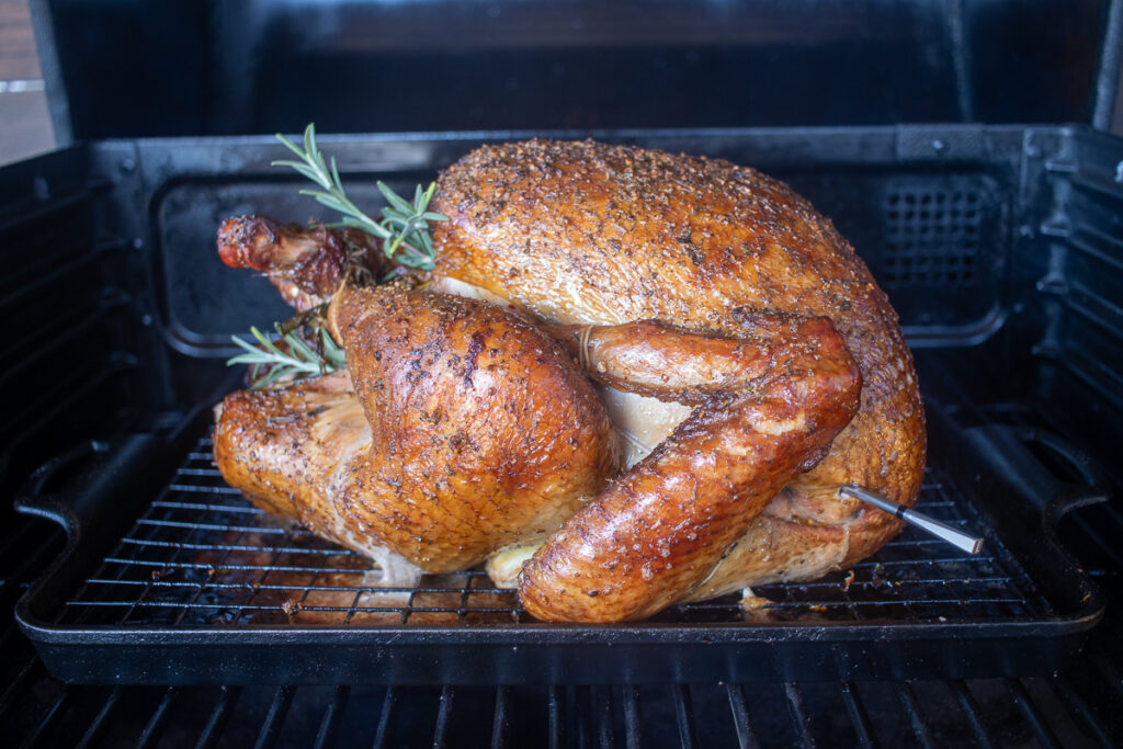 A perfectly smoked turkey resting on a wire rack inside a pellet grill, featuring crispy, golden-brown skin and garnished with sprigs of fresh rosemary, with a meat thermometer inserted for precise cooking.