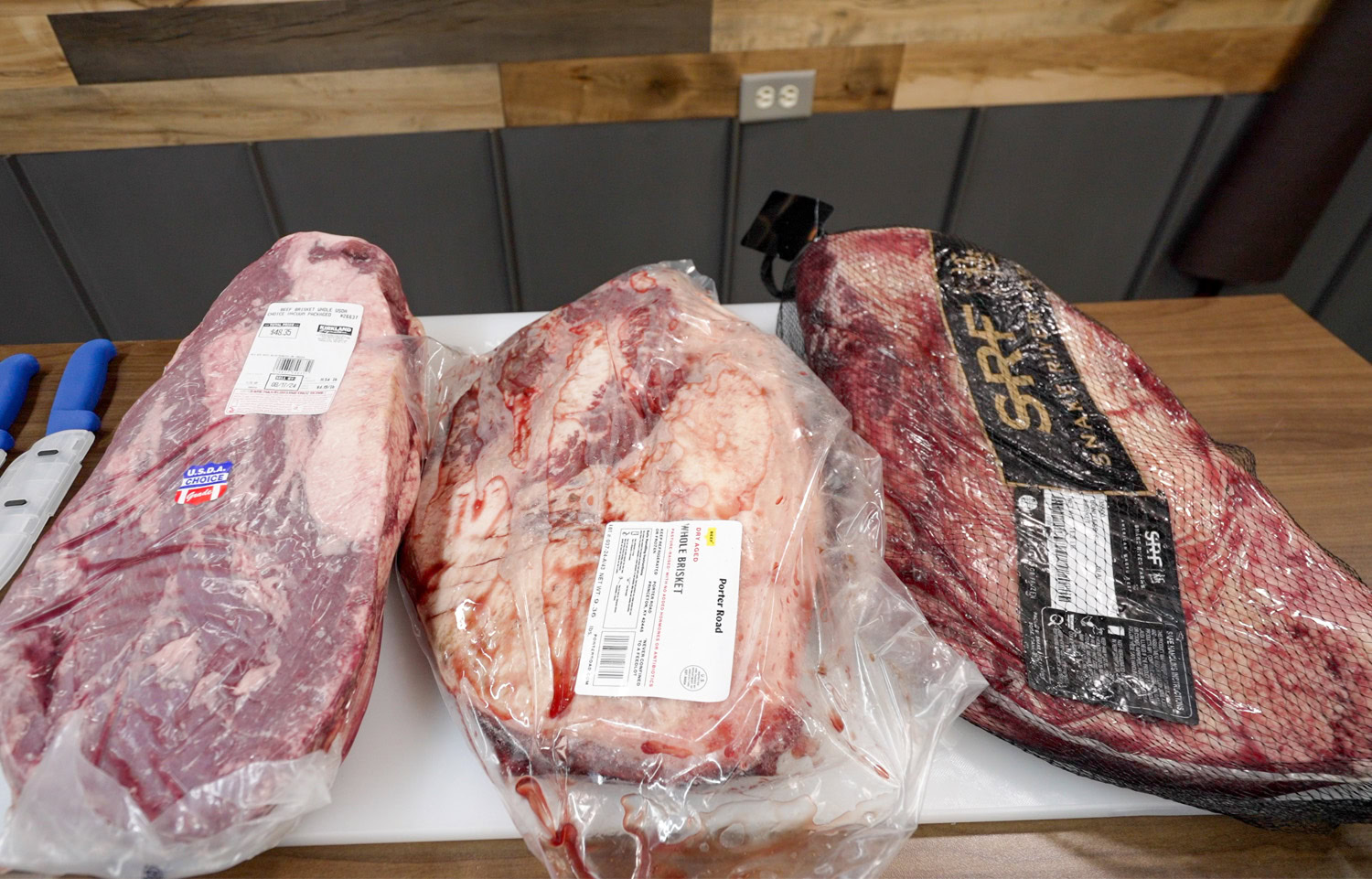 Three raw whole briskets in packaging displayed side by side on a butcher block—Costco USDA Choice, Porter Road, and Snake River Farms—used in a smoked brisket comparison test.