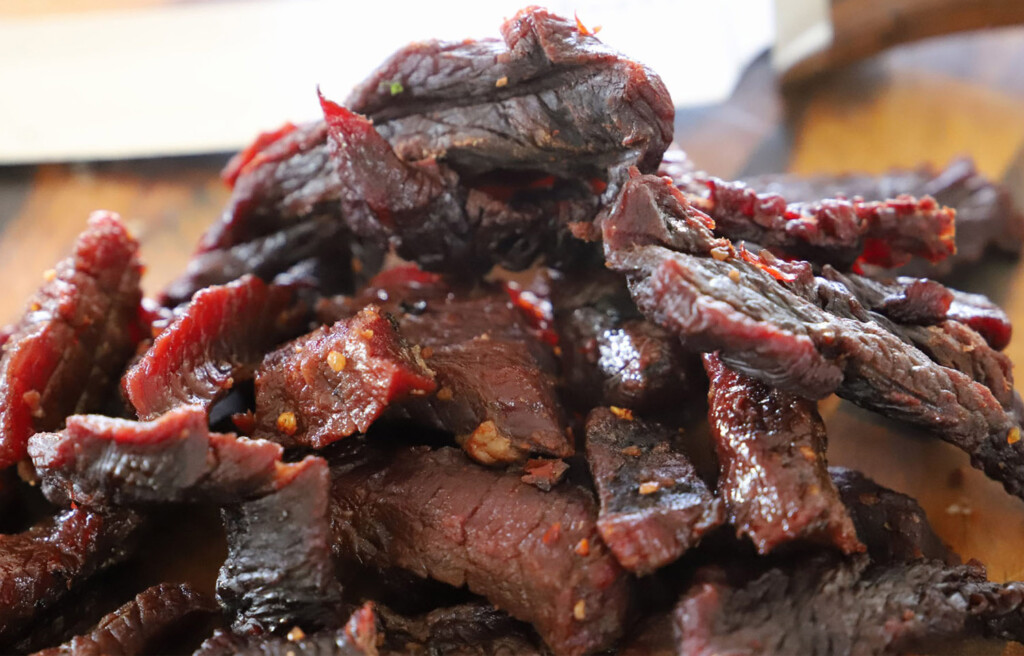 Close-up of sliced smoked beef jerky piled on a wooden cutting board, showing a dark, glossy exterior with a reddish interior and visible chili flakes.