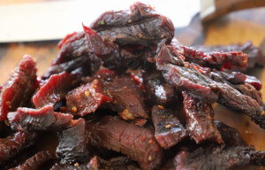 Close-up of sliced smoked beef jerky piled on a wooden cutting board, showing a dark, glossy exterior with a reddish interior and visible chili flakes.