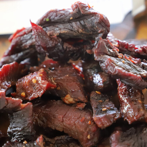 Close-up of sliced smoked beef jerky piled on a wooden cutting board, showing a dark, glossy exterior with a reddish interior and visible chili flakes.