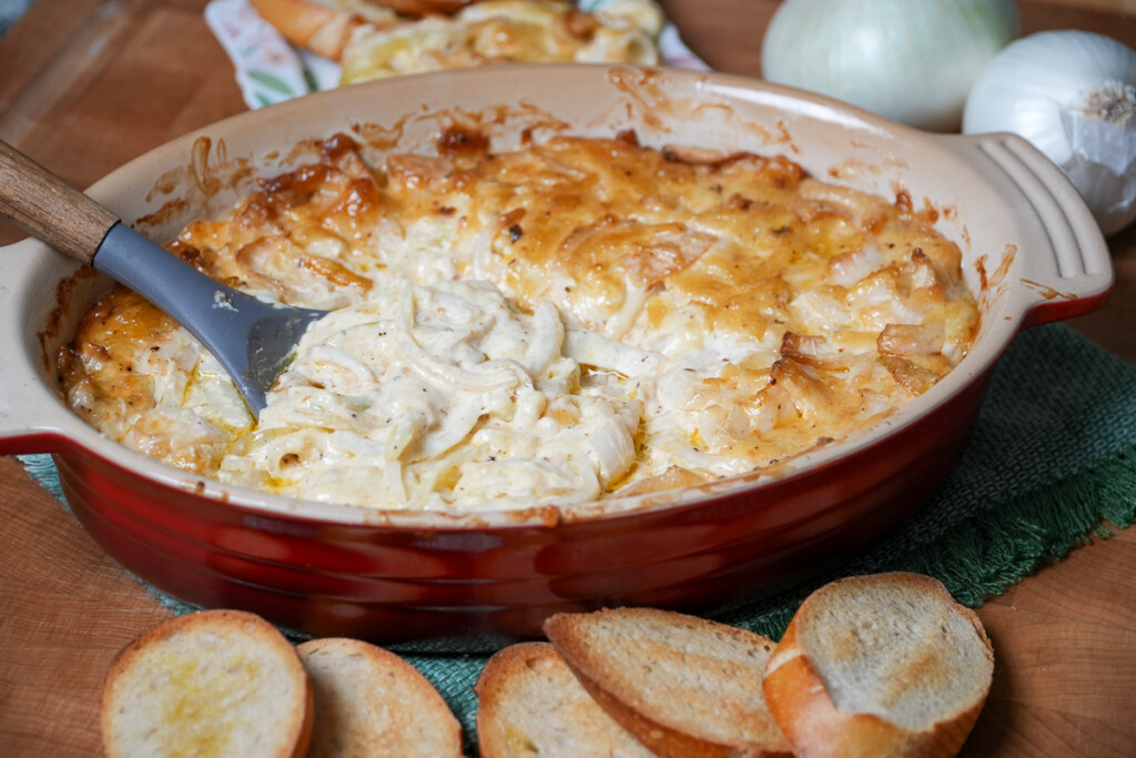 a spoon scooping some of the smoked onion dip out of the red casserole dish, with french bread slices in front