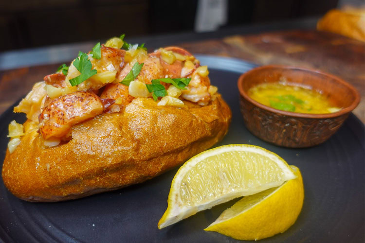 a lobster roll on a black plate with sliced lemon and a bowl of melted garlic butter