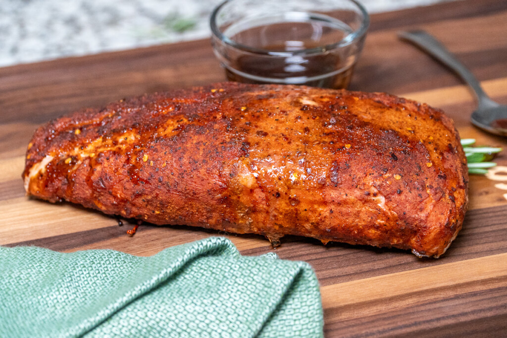 a cooked piece of pork loin on a wood board with a green napkin in front 