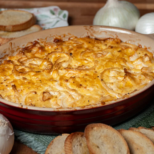 Bubbling smoked hot onion dip in a red ceramic dish, topped with golden-brown melted cheese, surrounded by white onions and toasted bread slices on a wooden table.