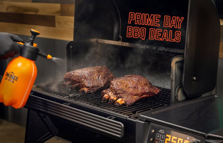 Two racks of smoked beef ribs being spritzed inside a Traeger pellet grill, with bold text reading ‘Prime Day BBQ Deals’ in orange outline over the open grill lid