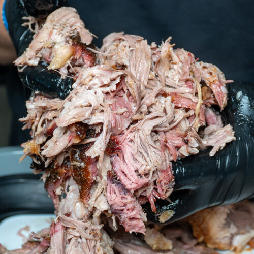 Close-up of shredded smoked pork being pulled apart by a person wearing black gloves. The meat is tender with a pink smoke ring and crispy bark pieces, indicating it was slow-cooked on a smoker or pellet grill.