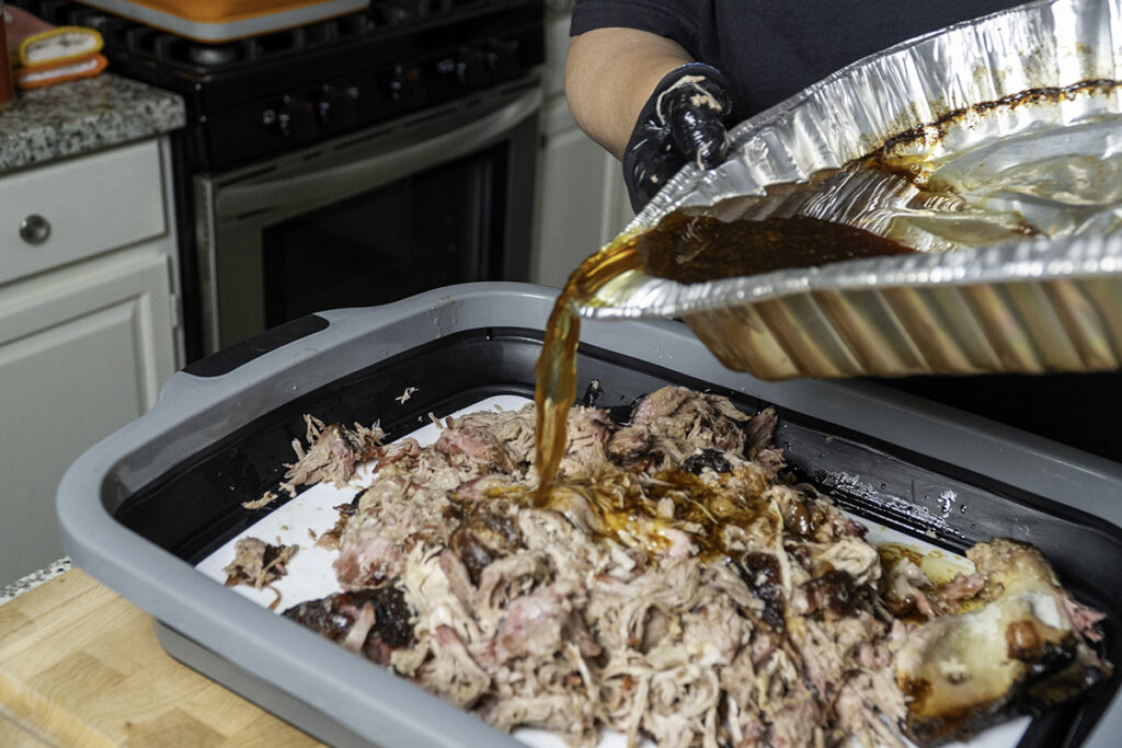 A person wearing black gloves pours flavorful cooking juices from a foil pan onto freshly shredded pulled pork in a large roasting tray. The pork has a mix of crispy bark and tender, juicy meat.