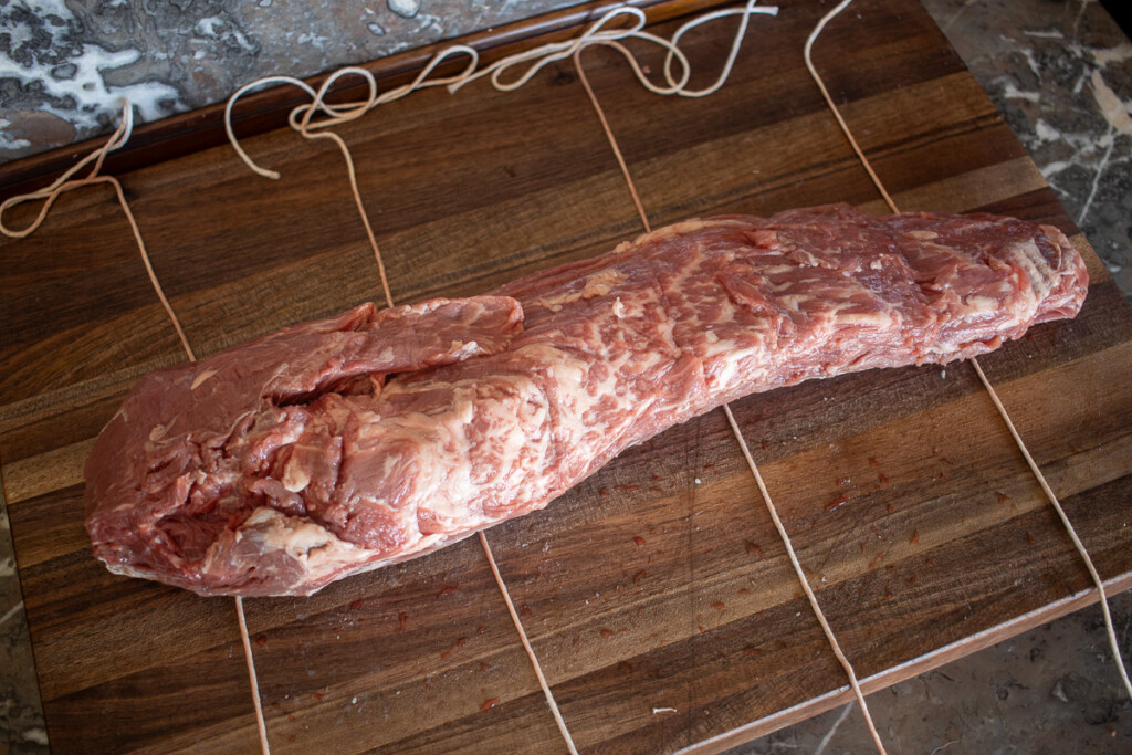 four strings of butchers twin on a wooden board with a raw beef tenderloin lying on them.