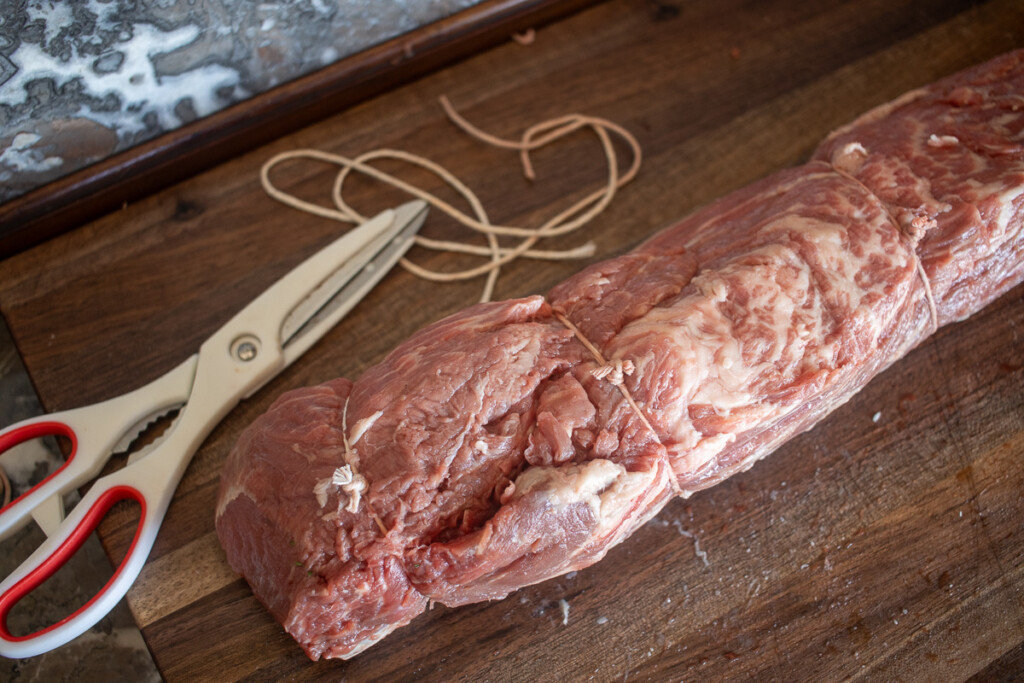 a raw beef tenderloin tied with butchers twine, on a wooden board with scissors and twine beside it