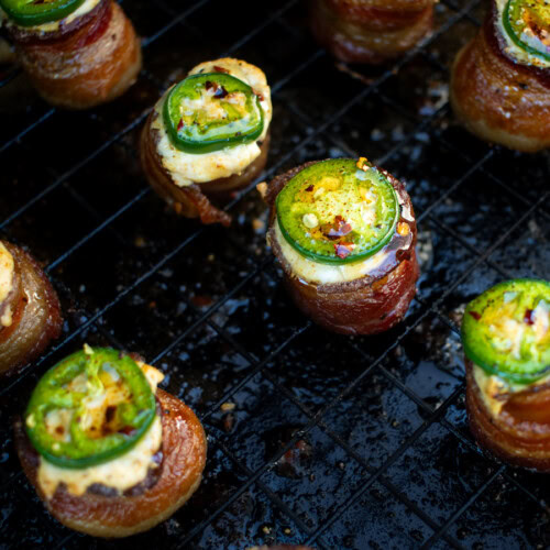 Close-up of smoked pig shots on a wire rack, showing crispy bacon-wrapped sausage bites filled with melted cream cheese and topped with sliced jalapeños and red pepper flakes.