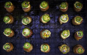 Overhead view of smoked pig shots on a wire rack, featuring crispy bacon-wrapped sausage cups filled with golden cream cheese, topped with sliced jalapeño and sweet BBQ seasoning.