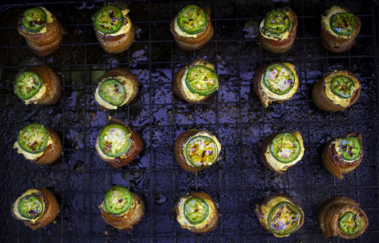 Overhead view of smoked pig shots on a wire rack, featuring crispy bacon-wrapped sausage cups filled with golden cream cheese, topped with sliced jalapeño and sweet BBQ seasoning.