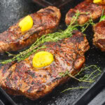 Four New York strip steaks searing on a Blackstone griddle, topped with melting ghee and surrounded by sprigs of rosemary and thyme.