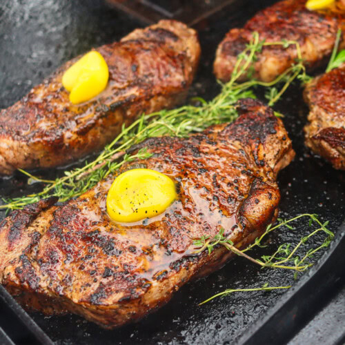 Four New York strip steaks searing on a Blackstone griddle, topped with melting ghee and surrounded by sprigs of rosemary and thyme.