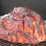 Close-up of a smoked ham on a wire rack, with caramelized glaze and deep bark from the smoker. Slits cut into the ham reveal tender, juicy meat underneath a sticky, flavor-packed crust.