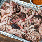 Tray of smoked pulled pork with dark bark and pink smoke ring, served in a foil pan on a wooden table with a bowl of dry rub in the background.
