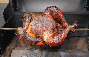 The image shows a well-cooked, crispy, and browned turkey mounted on a rod in a roasting oven. This process suggests an effective rotispliance technique, possibly involving a smoker given the rich coloring and texture of the turkey's skin.