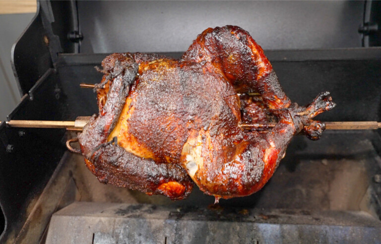 The image shows a well-cooked, crispy, and browned turkey mounted on a rod in a roasting oven. This process suggests an effective rotispliance technique, possibly involving a smoker given the rich coloring and texture of the turkey's skin.