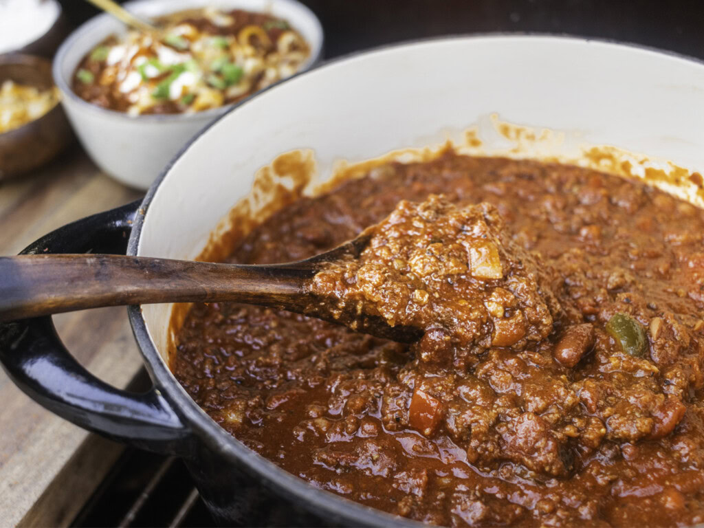 Close-up of smoked over the top chili in a Dutch oven, with a wooden spoon lifting a hearty scoop of chunky beef and beans.