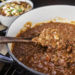 Close-up of smoked over the top chili in a Dutch oven, with a wooden spoon lifting a hearty scoop of chunky beef and beans.