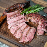 Sliced smoked tomahawk steak fanned out on a wooden cutting board, showing a pink medium-rare interior and charred crust, with the bone intact and garnished with fresh rosemary and a knife in the background.