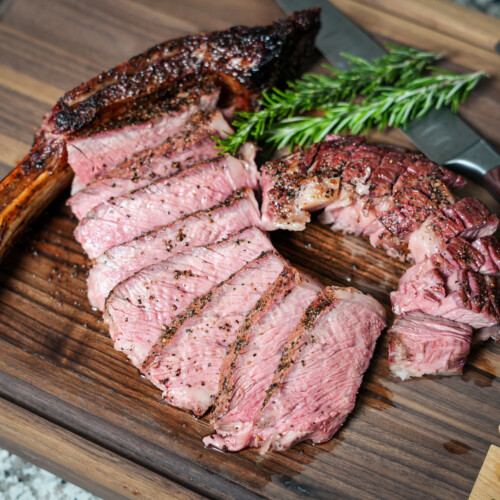 Sliced smoked tomahawk steak fanned out on a wooden cutting board, showing a pink medium-rare interior and charred crust, with the bone intact and garnished with fresh rosemary and a knife in the background.