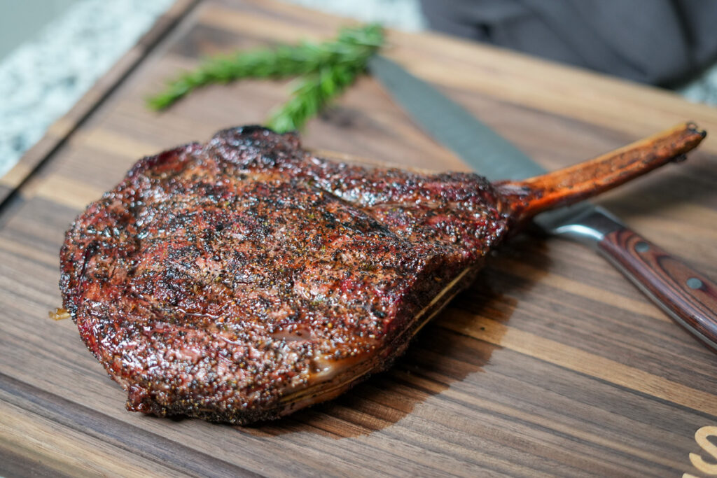 Reverse seared tomahawk steak resting on a wooden cutting board, showcasing a dark, crusty exterior and long bone, with a knife and rosemary in the background.