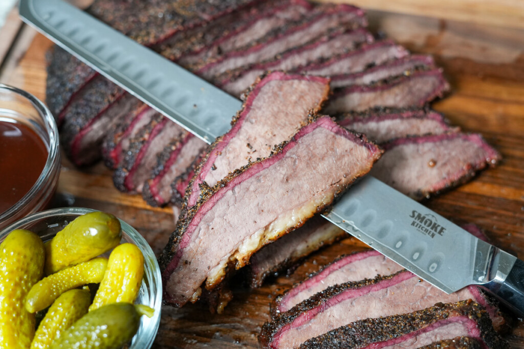 a sliced whole brisket in the background and two slices of brisket balancing on a knife