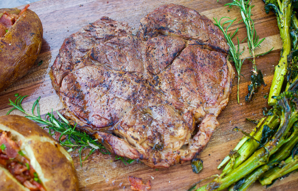 A heart-shaped ribeye steak with a beautifully seared crust sits on a wooden cutting board, surrounded by fresh rosemary, roasted asparagus, and a baked potato.