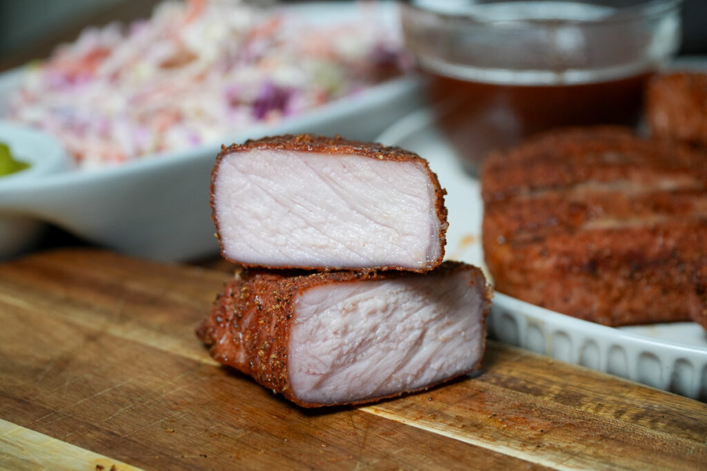 A close-up of perfectly smoked pork chops sliced in half, revealing a juicy, tender interior with a beautifully seasoned crust. In the background, coleslaw and barbecue sauce are visible, highlighting the perfect barbecue pairing.