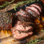 Juicy slices of smoked beef tenderloin with a flavorful herb crust, presented on a wooden cutting board garnished with sprigs of fresh rosemary