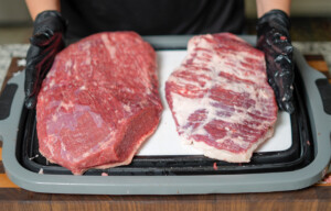 Two separated brisket muscles—the flat on the left and the point on the right—resting on a cutting board. A person wearing black nitrile gloves holds each piece, showcasing the difference in texture and marbling between the lean flat and the fattier point.