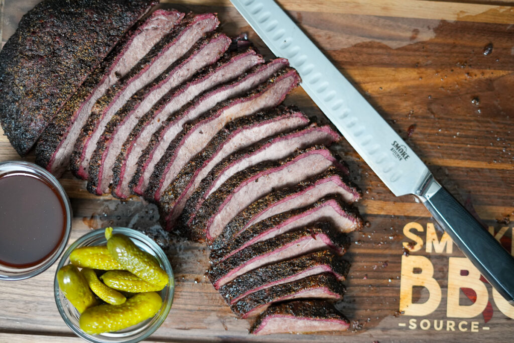 A sliced smoked brisket flat on a wooden cutting board, showing a dark crust, pink smoke ring, and juicy interior. A carving knife, pickles, and a bowl of barbecue sauce sit alongside."Let me know if you'd like any tweaks!