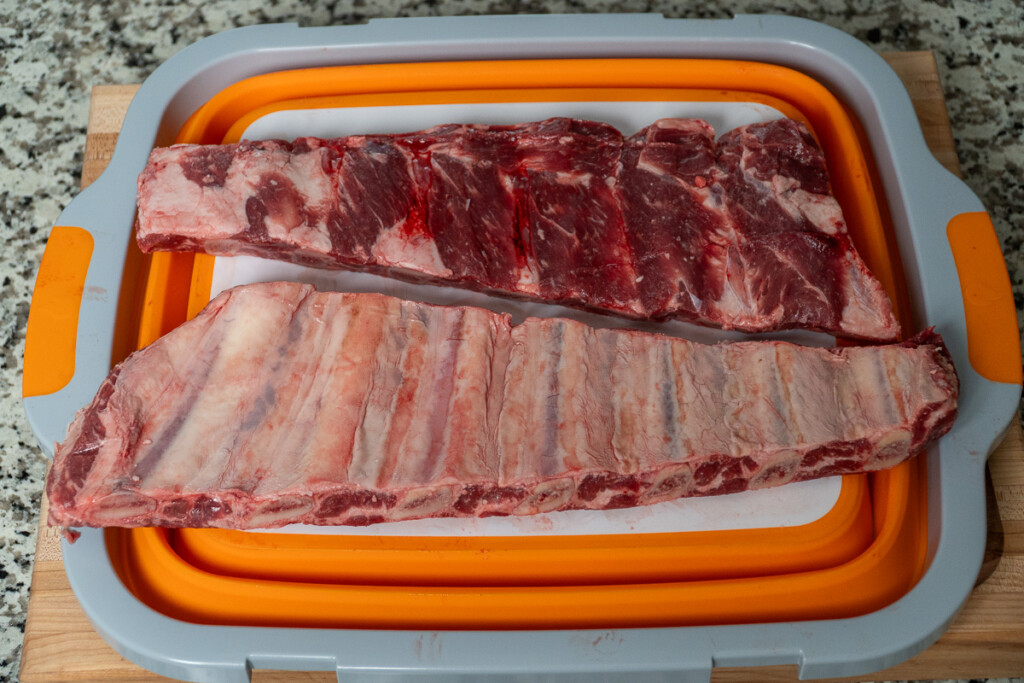 two slabs of beef back ribs, one showing front, one showing back on a orange and whte board