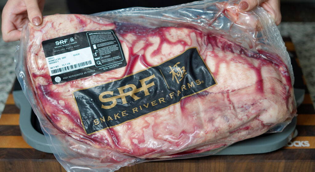 A person holds a vacuum-sealed Snake River Farms American Wagyu brisket, labeled as "Gold Grade." The marbled beef is visible through the packaging, showcasing its high-quality fat distribution.