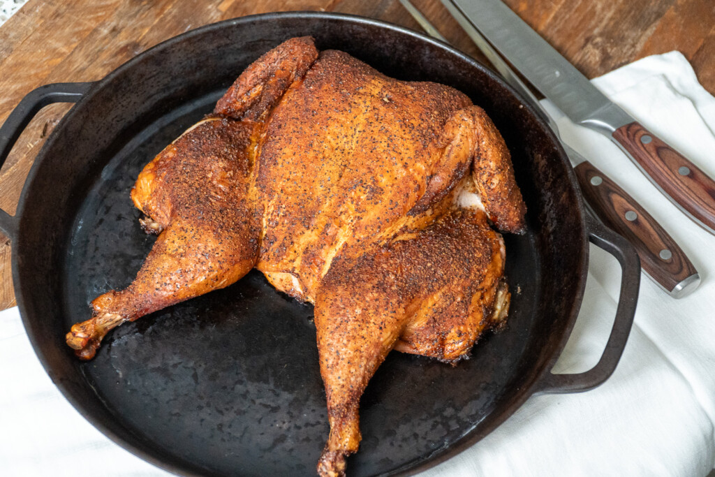 Spatchcocked smoked chicken seasoned with dry rub, resting in a cast iron skillet with carving knives nearby.