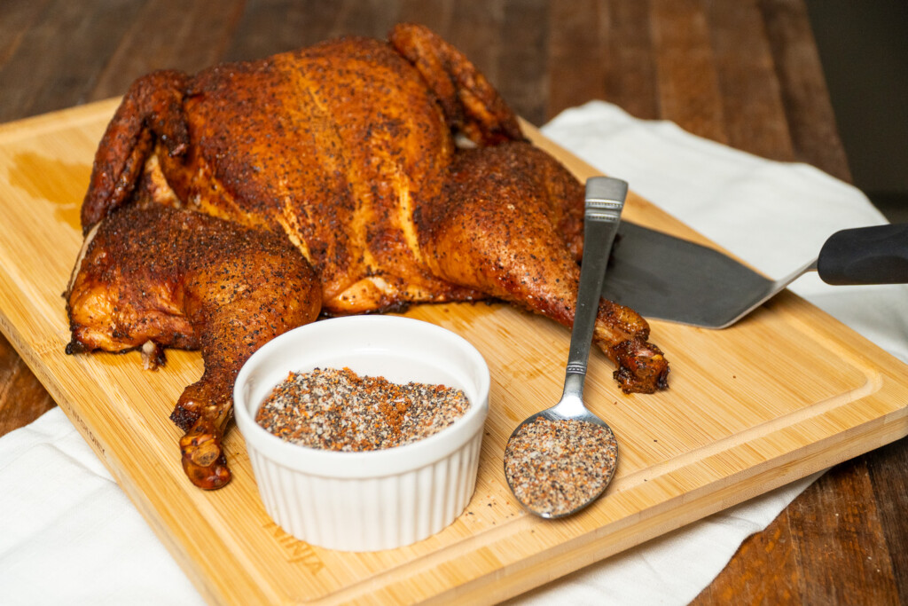 a cooked roast spatchcock chicken with a bowl of chicken rub and a spoonful of chicken rub all on a wooden board