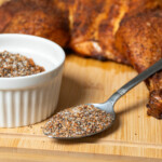 Close-up of a smoked chicken rub in a white ramekin and spoon, with seasoned smoked chicken in the background on a wooden cutting board.